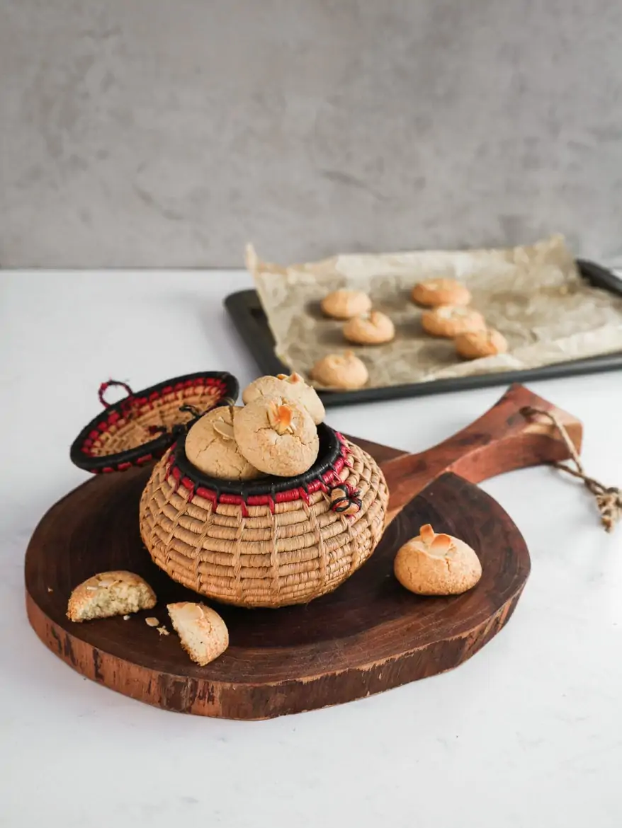 Angle shot of a traditional round basket filled to the brim with almond flour cookies topped with sliced almonds on a round wooden board with a broken cookie. There is also a baking sheet in the background with cookies.