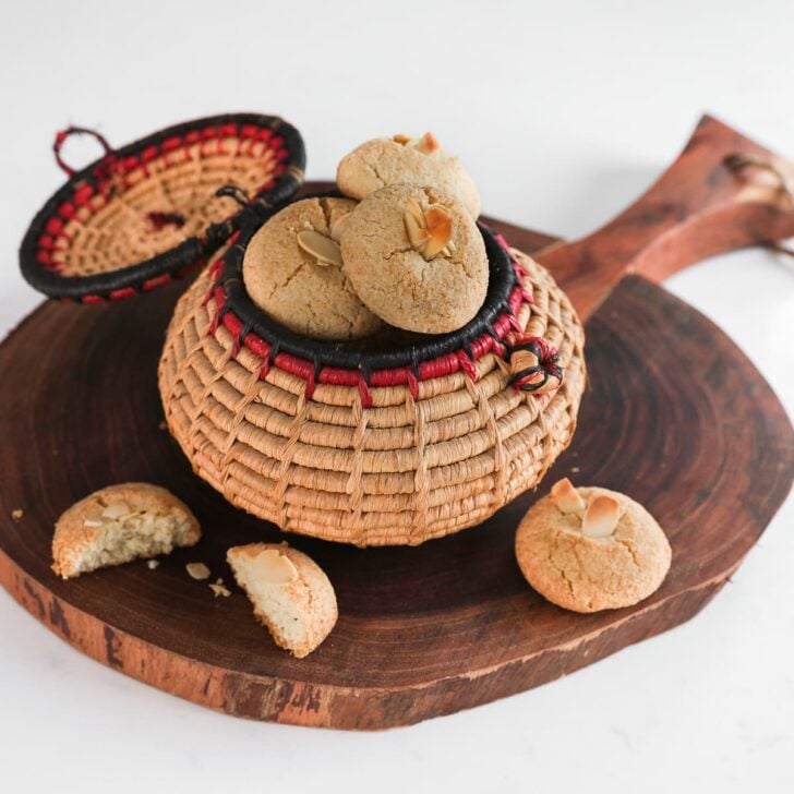 a traditional round basket filled to the brim with almond flour cookies topped with sliced almonds on a round wooden board