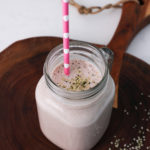 a glass jar of homemade protein shake that's pink in colour and topped with hemp hearts with a pink polka dotted straw inside.