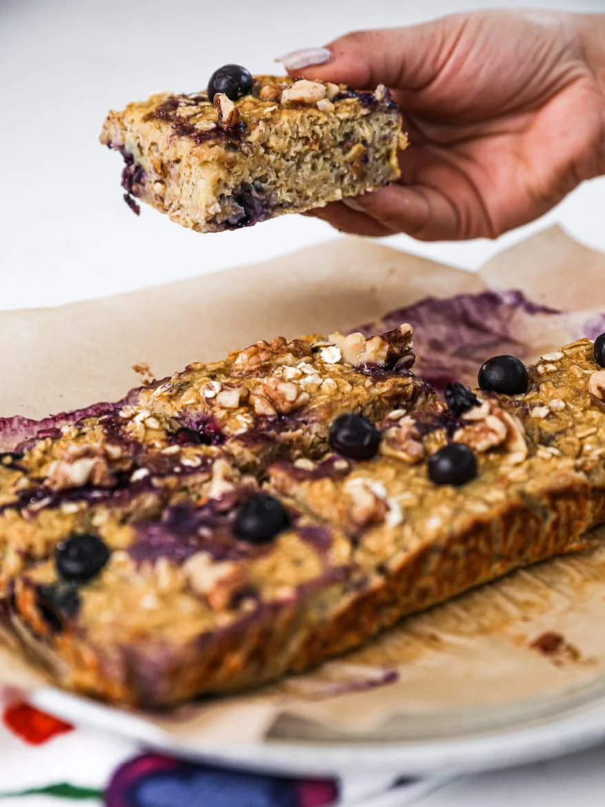 someone holding a baked oatmeal square over a plate with five other squares topped with blueberries