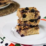 Baked oatmeal squares topped with walnuts and blueberries staked on top of one another on a white plate.