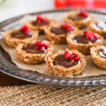 A close up shot of a round tray holding oat cup cookies that are filled with peanut butter and chocolate and topped with pomegranate kernels.