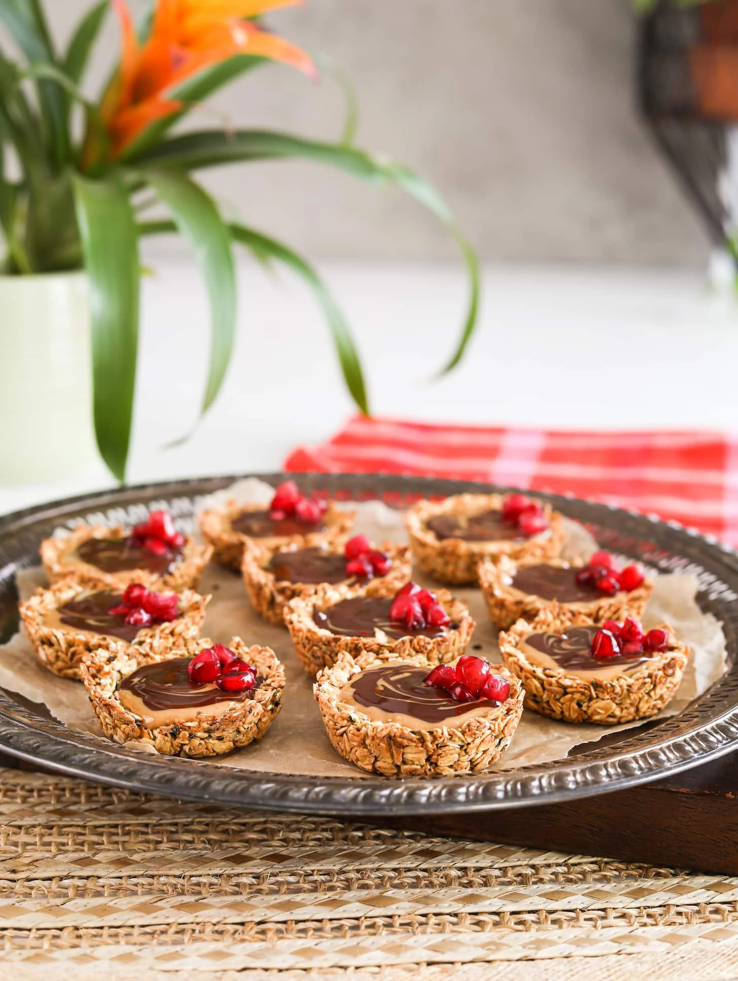 A round tray holding oat cup cookies that are filled with peanut butter and chocolate and topped with pomegranate kernels. There is a plant in the distance.