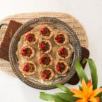 A round tray holding oat cup cookies that are filled with peanut butter and chocolate and topped with pomegranate kernels. It's placed on a wooden board.