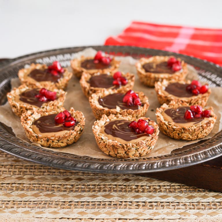 A round tray holding oat cup cookies that are filled with peanut butter and chocolate and topped with pomegranate kernels.