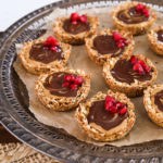 An angled overhead shot of a round tray holding oat cup cookies that are filled with peanut butter and chocolate and topped with pomegranate kernels.