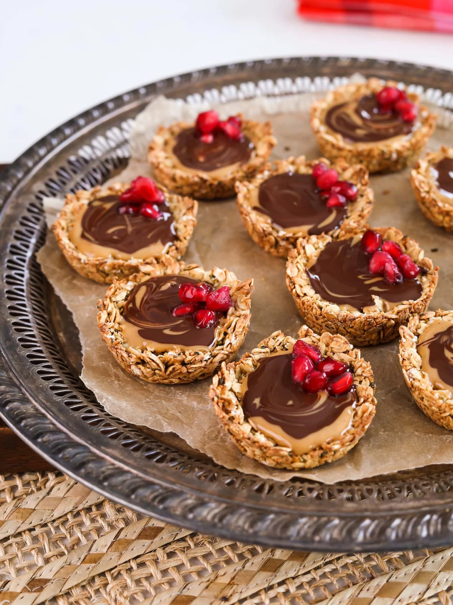 An angled overhead shot of a round tray holding oat cup cookies that are filled with peanut butter and chocolate and topped with pomegranate kernels.