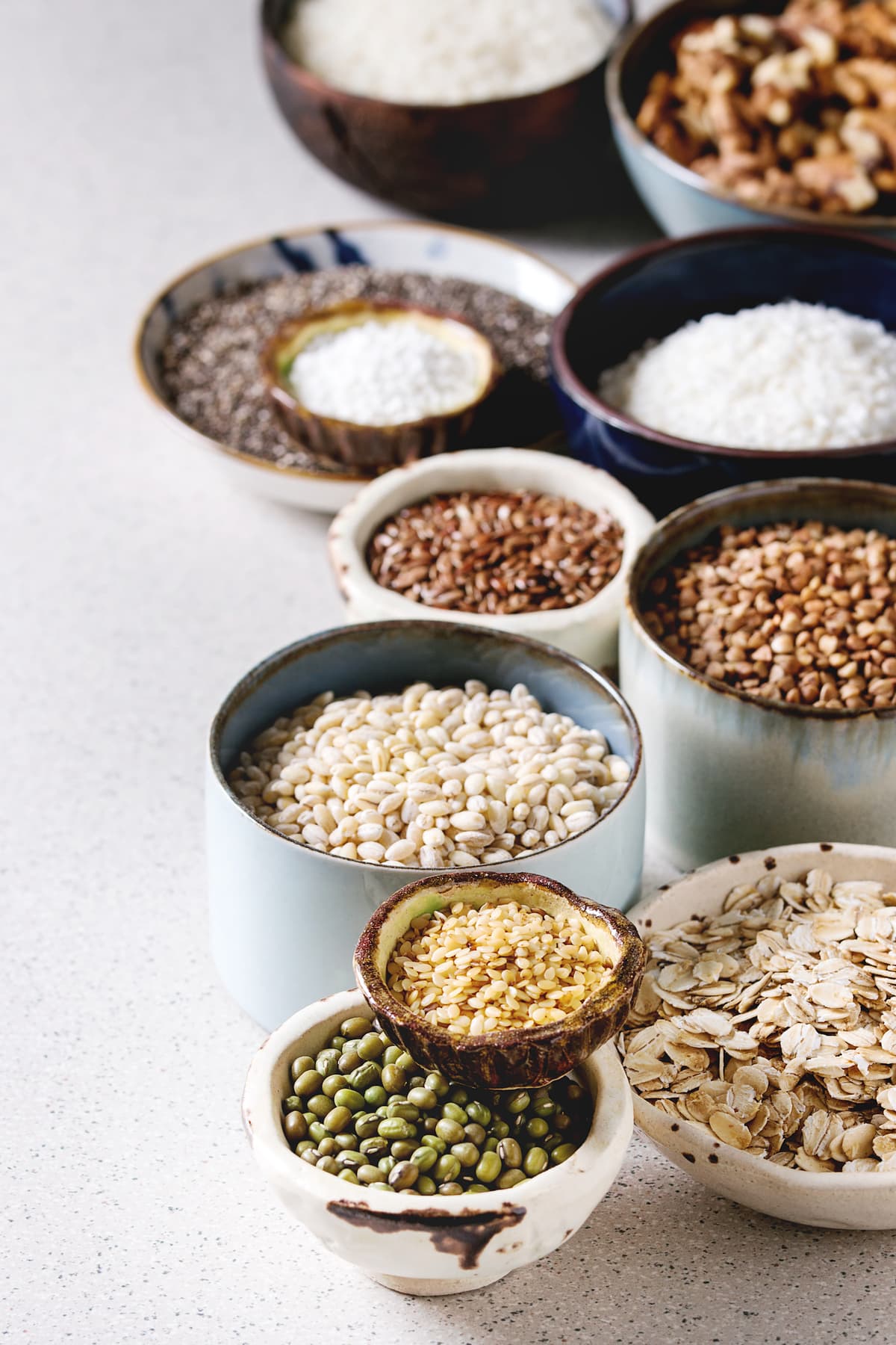 Variety of raw uncooked grains superfood cereal chia seeds, linen, sesame, mung bean, walnuts, tapioca, wheat, buckwheat, oatmeal, coconut, rice in ceramic bowls over grey spotted background.