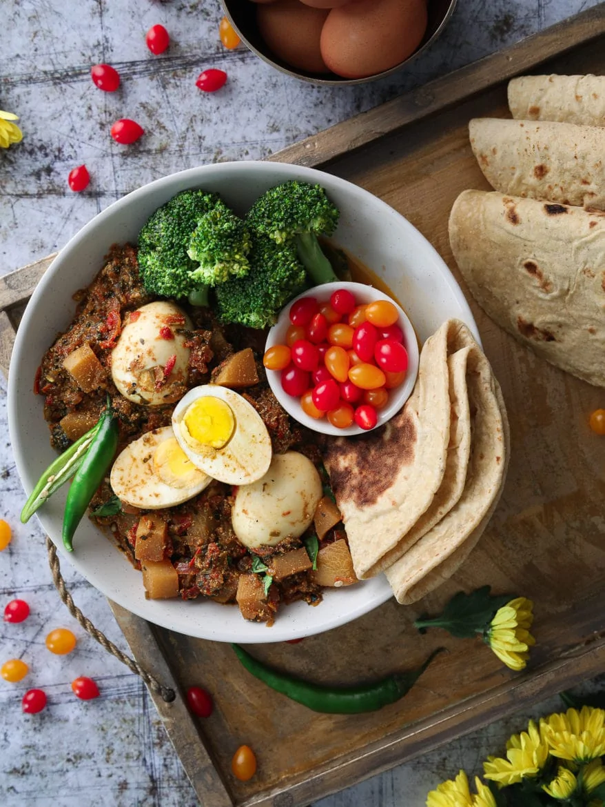 a bowl of egg curry with turnips with a side of broccoli, baby tomatoes and roti presented on a tray with folded rotis.