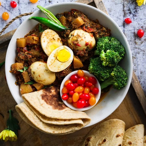 a bowl of egg curry with turnips with a side of broccoli, baby tomatoes and roti presented on a tray.