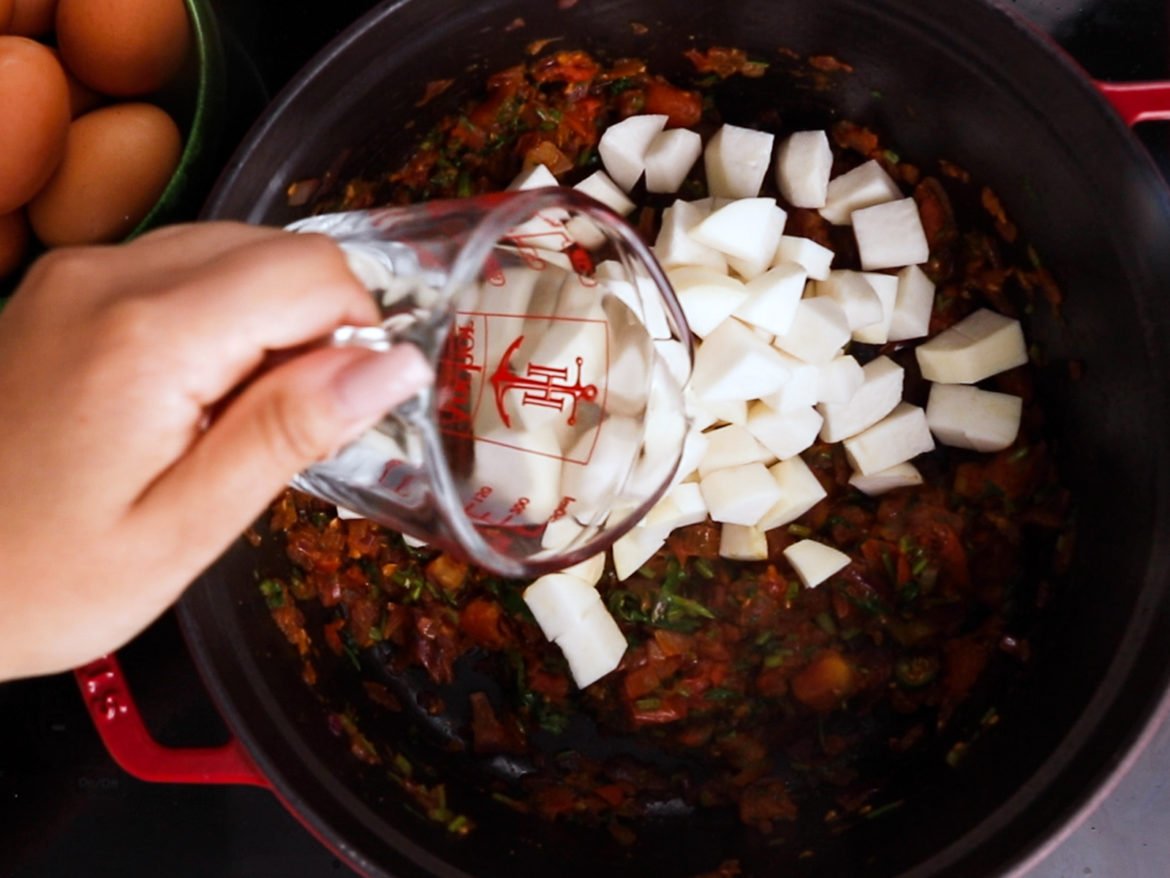 a hand pouring a jug of water into a cooking pot with turnip cubes and cooked tomatoes.