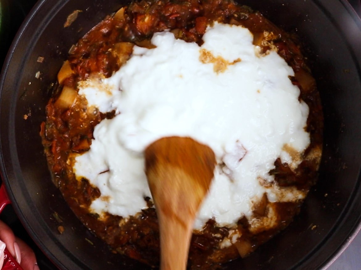a cooking pot with yogurt over a red curry sauce with a wooden spoon submerged in the yogurt