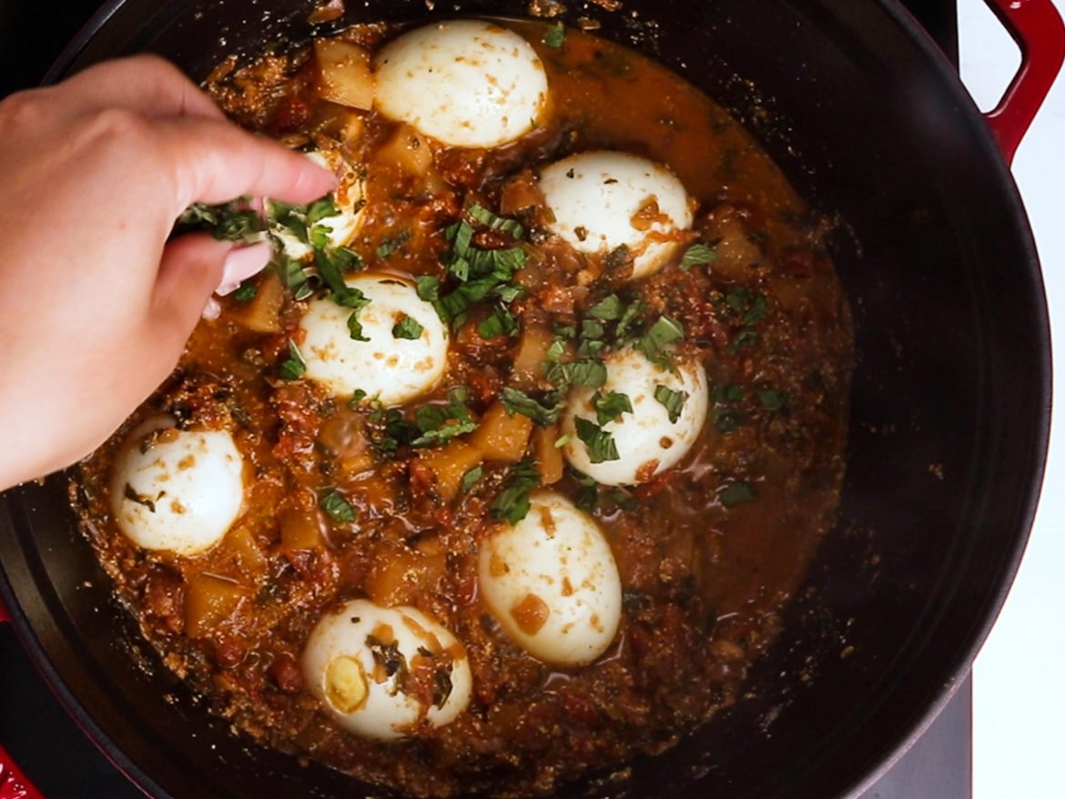 a hand sprinkling green herbs over egg curry with turnips in a cooking pot.