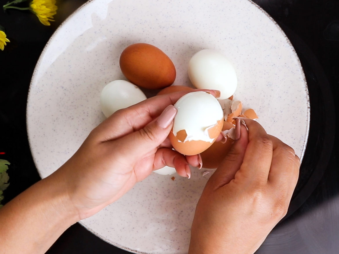 a person's hands showing peeling of a boiled egg over a plate of eggs