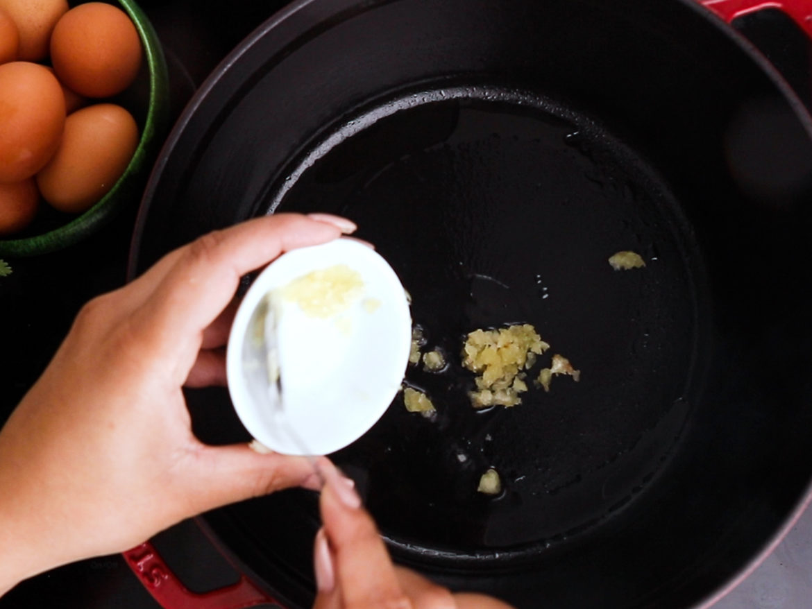a hand holding a ramekin adding garlic with a spoon to a cooking pot with oil.