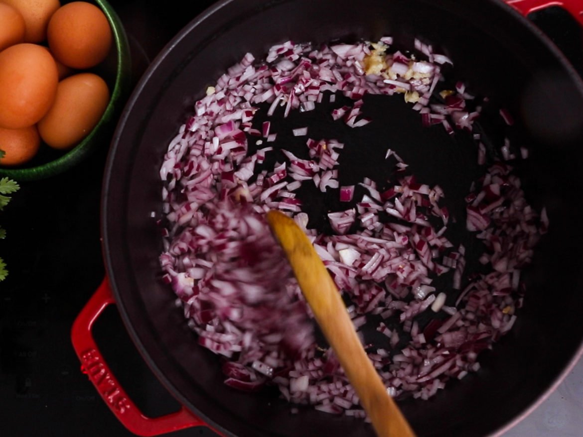 a cooking pot with a wooden spoon inside and chopped red onion, with a bowl of eggs next to it.