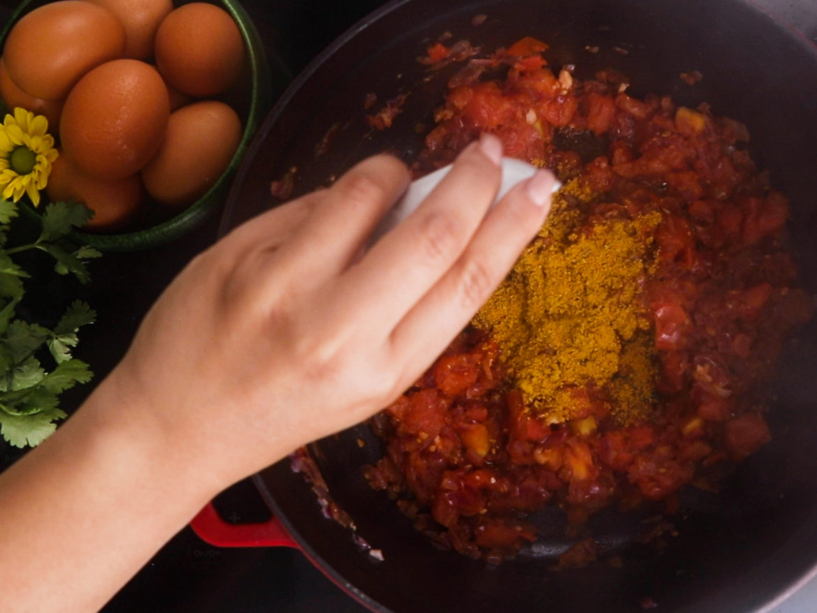 a hand adding a yellow spice from a small ramekin to a cooking pot with tomatoes. A bowl of eggs with a yellow flower seen next to the pot.