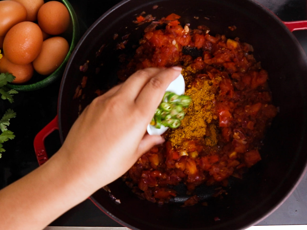 a hand holding a ramekin of chopped green chillies and adding these to a cooking pot with cooked tomatoes and a yellow spice. A bowl of eggs are displayed next to the pot.