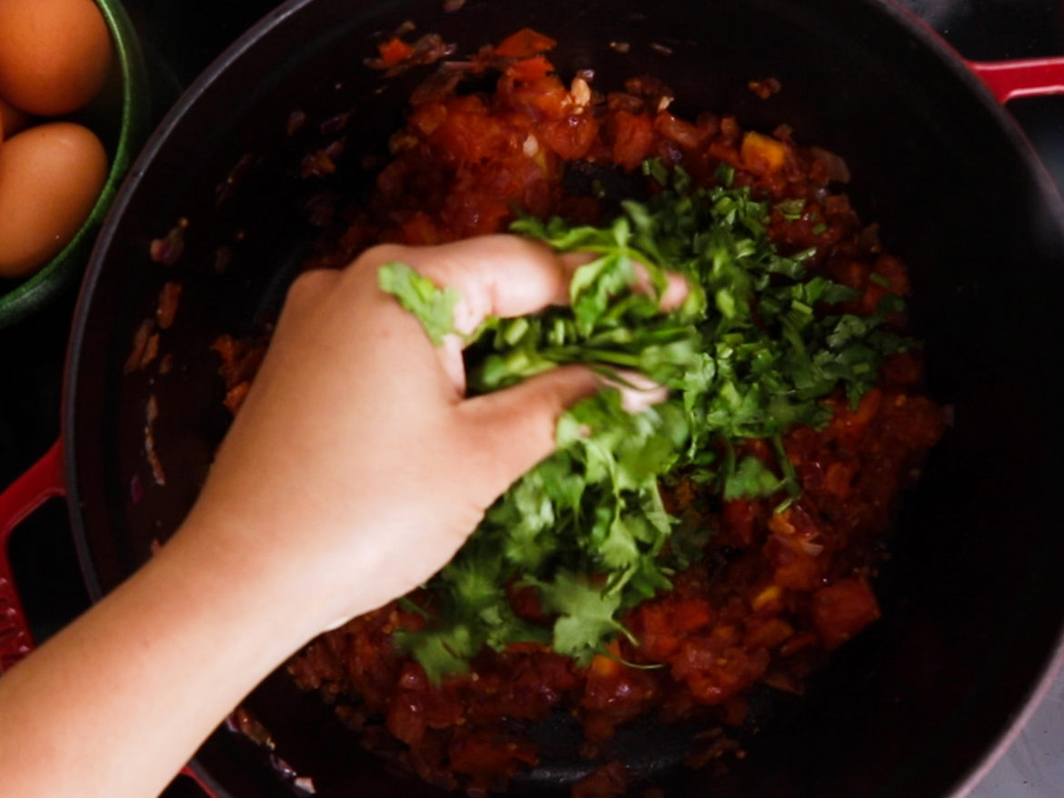 a hand adding chopped herbs to a cooking pot with cooked tomatoes.