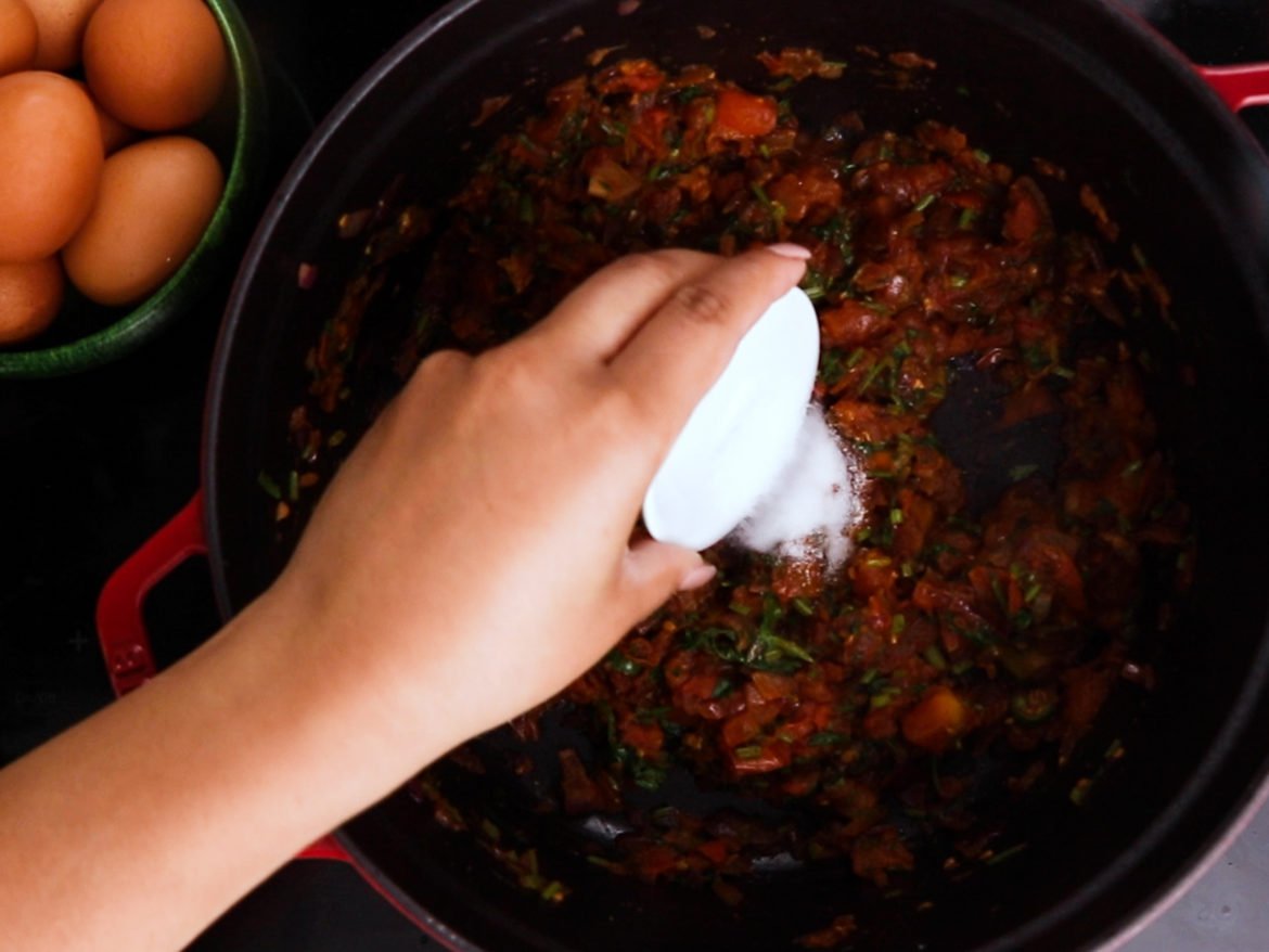 a hand holding a ramekin with salt and adding it to a cooking pot with cooked tomatoes and herbs