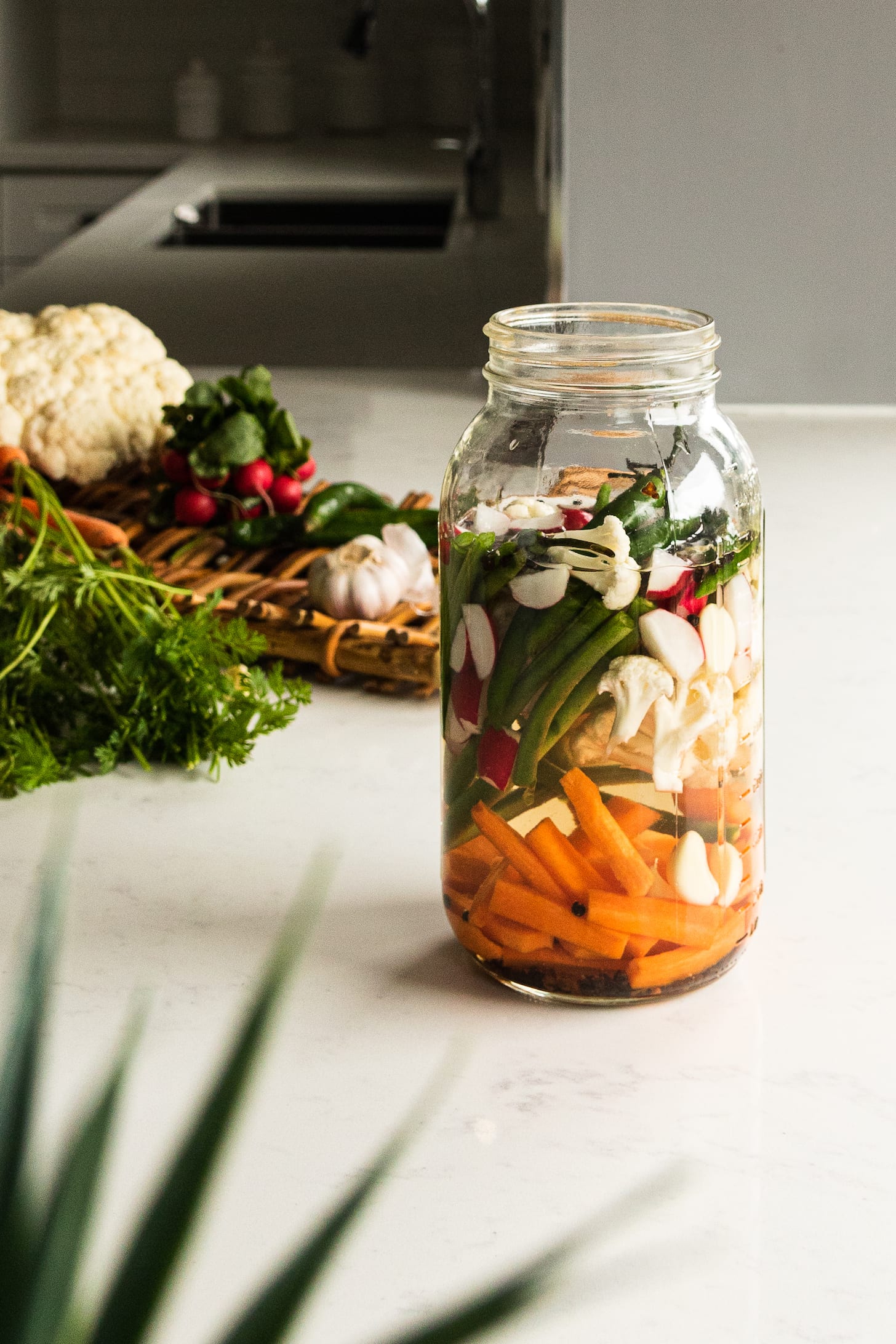 A jar of mixed vegetables in water on a countertop with fresh vegetables in the background.