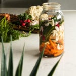 a jar of mixed vegetables in water with a plant in the foreground and vegetables in the background.