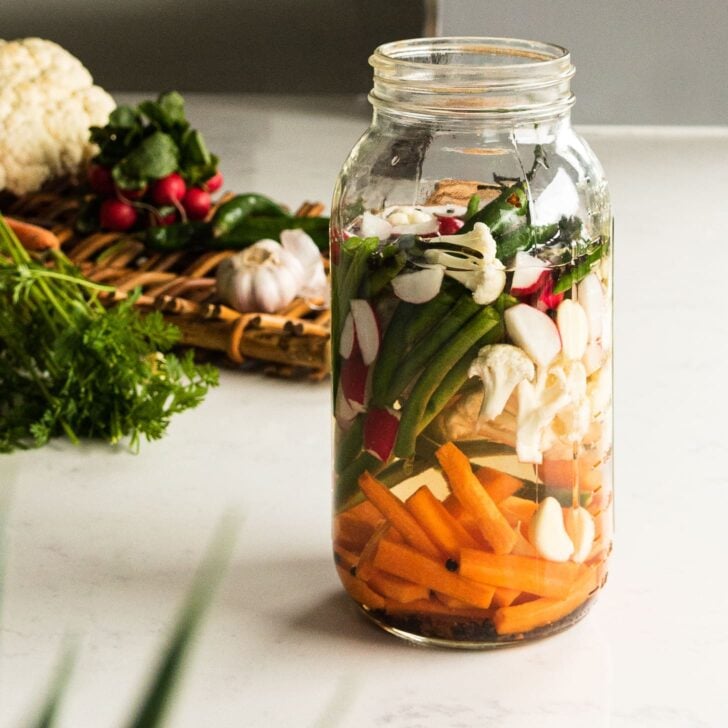 a close up image of a jar of mixed vegetables with water. Some vegetables in the background in a tray