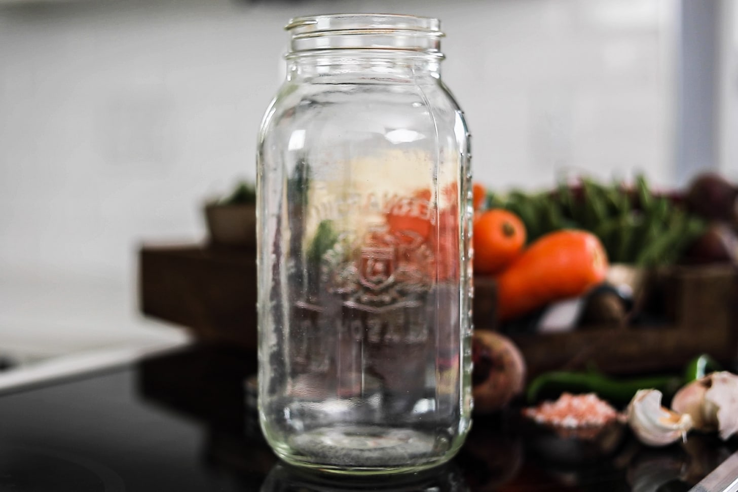 An empty mason jar with vegetables in a tray in the background.