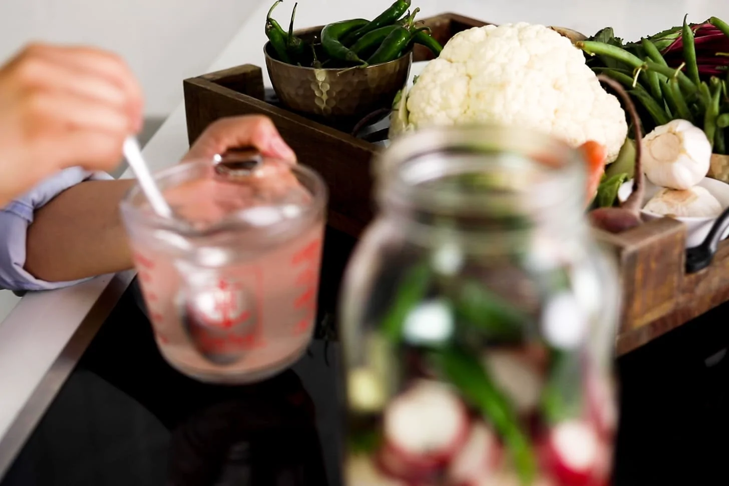 a jar of vegetables in the foreground with someone stirring a jug of water. There is also a tray of mixed vegetables in the background.