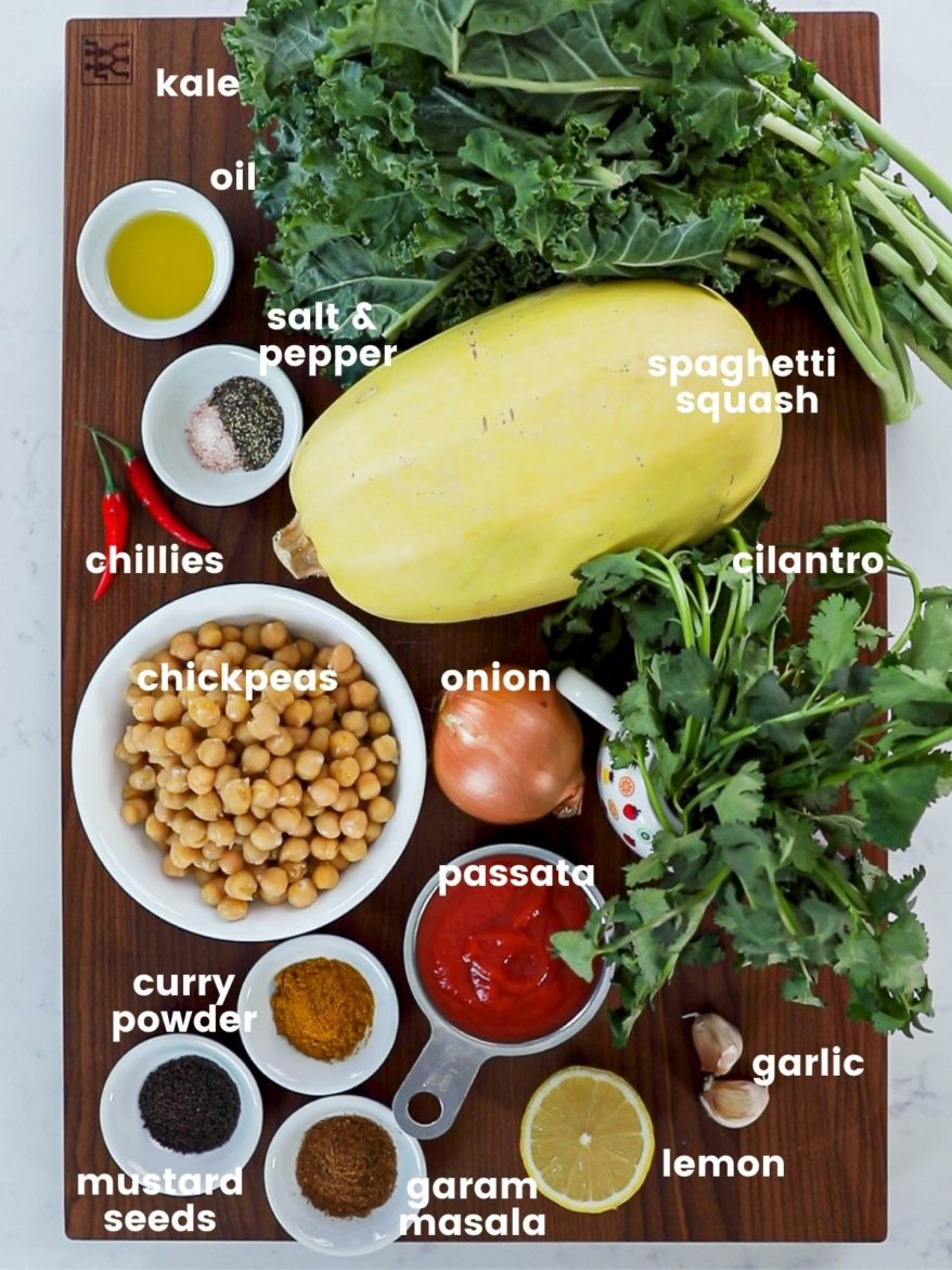 a rectangular wooden board with ingredients for chickpea curry arranged. It includes spaghetti squash, kale, bowl of chickpeas, ramekins of spices, oil, lemon and more.