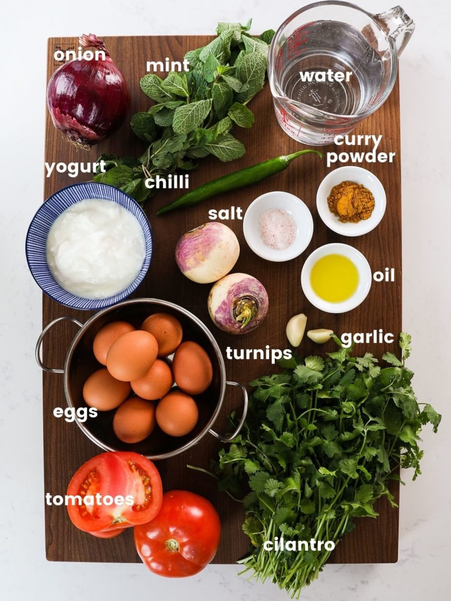 a rectangular wooden board displaying ingredients for egg curry with turnips. It includes eggs, yogurt, a jug of water, ramekins of spices, oil, as well as red onion and fresh herbs.