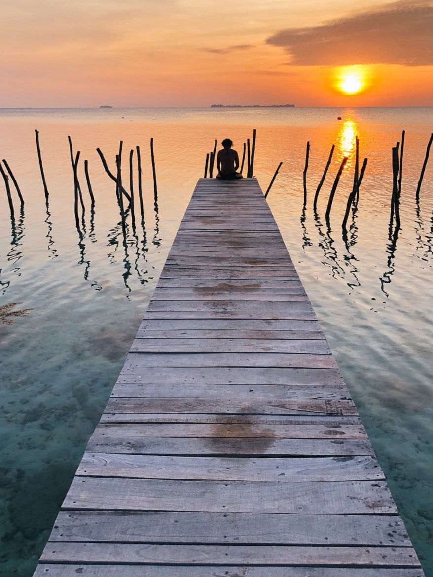 a person sitting at the end of a bridge overlooking water and a sunset.