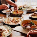table with an assortment of cooked dishes with several hands trying to help themselves to the food.