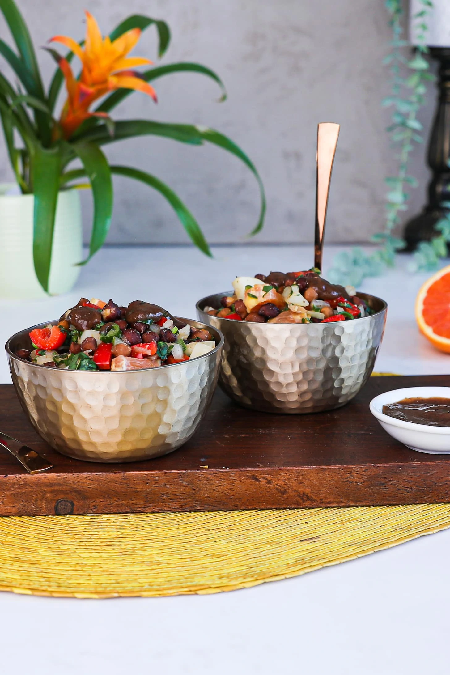 Two bowls of salad with chopped peppers, orange cubes, onion and herbs topped with a brown sauce placed on a wooden board.
