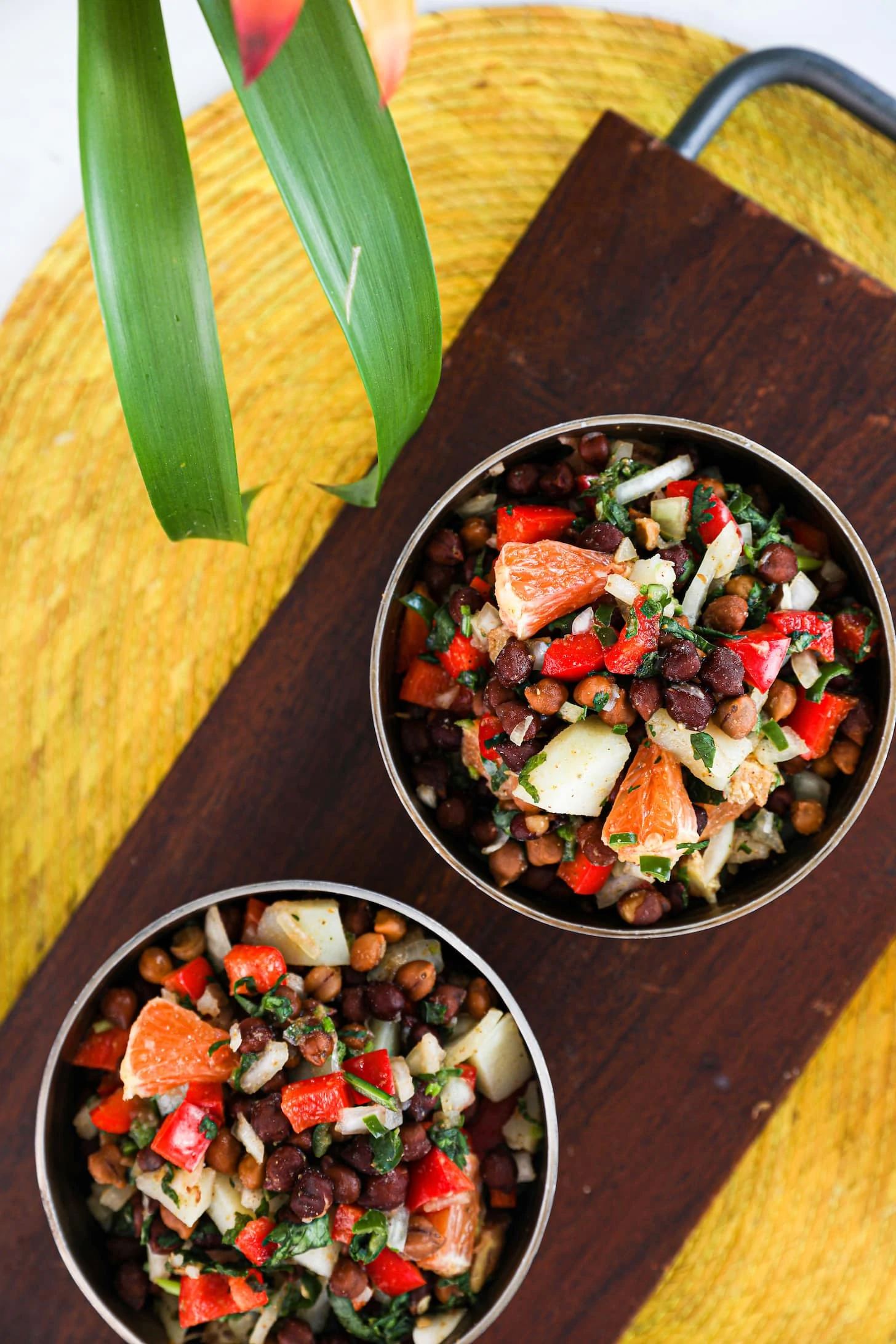 Overhead shot of two bowls of black chickpeas salad with orange cubes, peppers, potatoes and onion placed on a wooden board.
