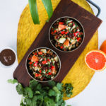 Overhead shot of two bowls of black chickpeas salad with orange cubes, peppers, potatoes and onion placed on a wooden board. Surrounded by a bunch of fresh herbs, orange halves, and a small bowl of brown sauce.