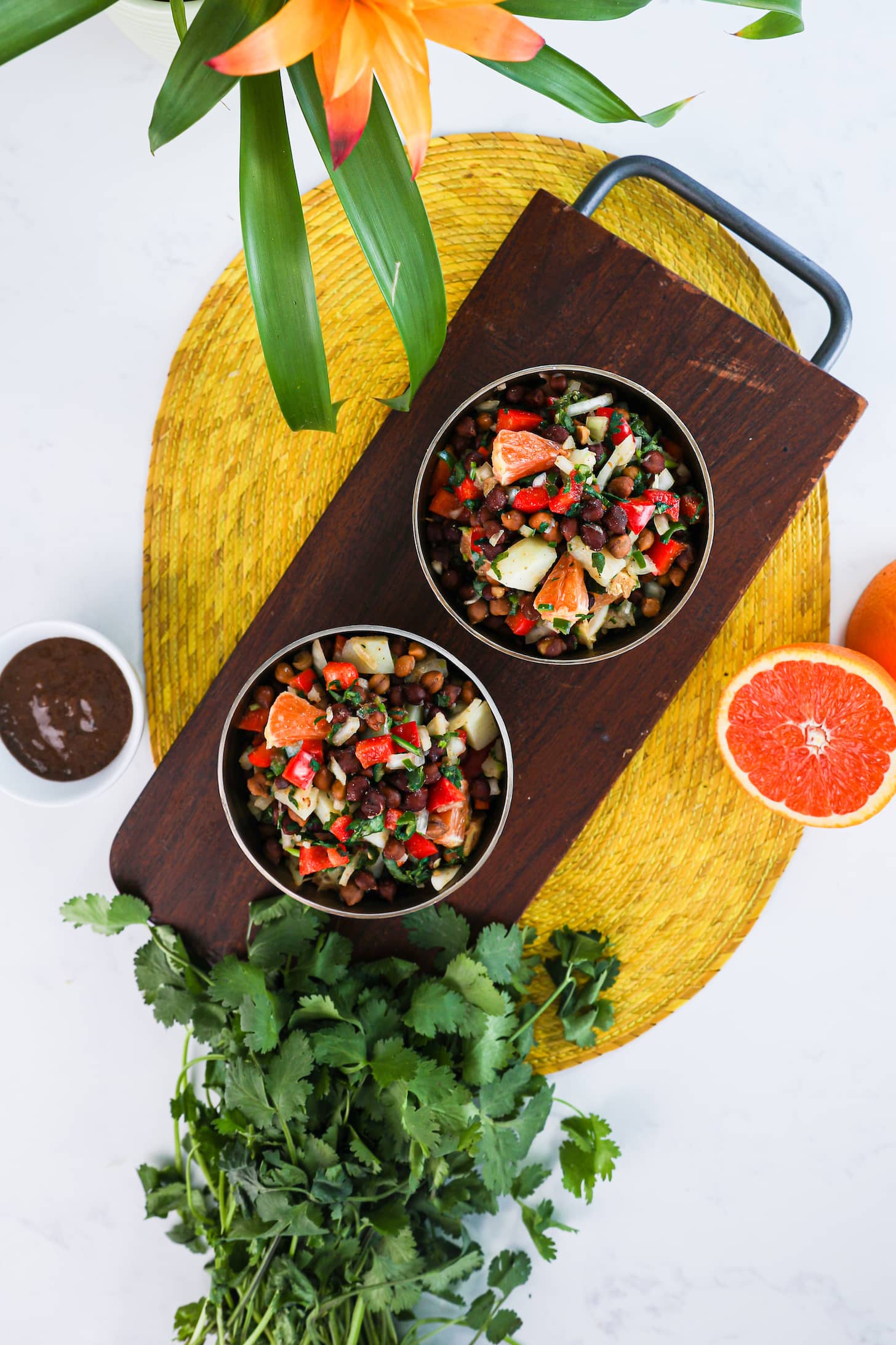 Overhead shot of two bowls of black chickpeas salad with orange cubes, peppers, potatoes and onion placed on a wooden board. Surrounded by a bunch of fresh herbs, orange halves, and a small bowl of brown sauce.