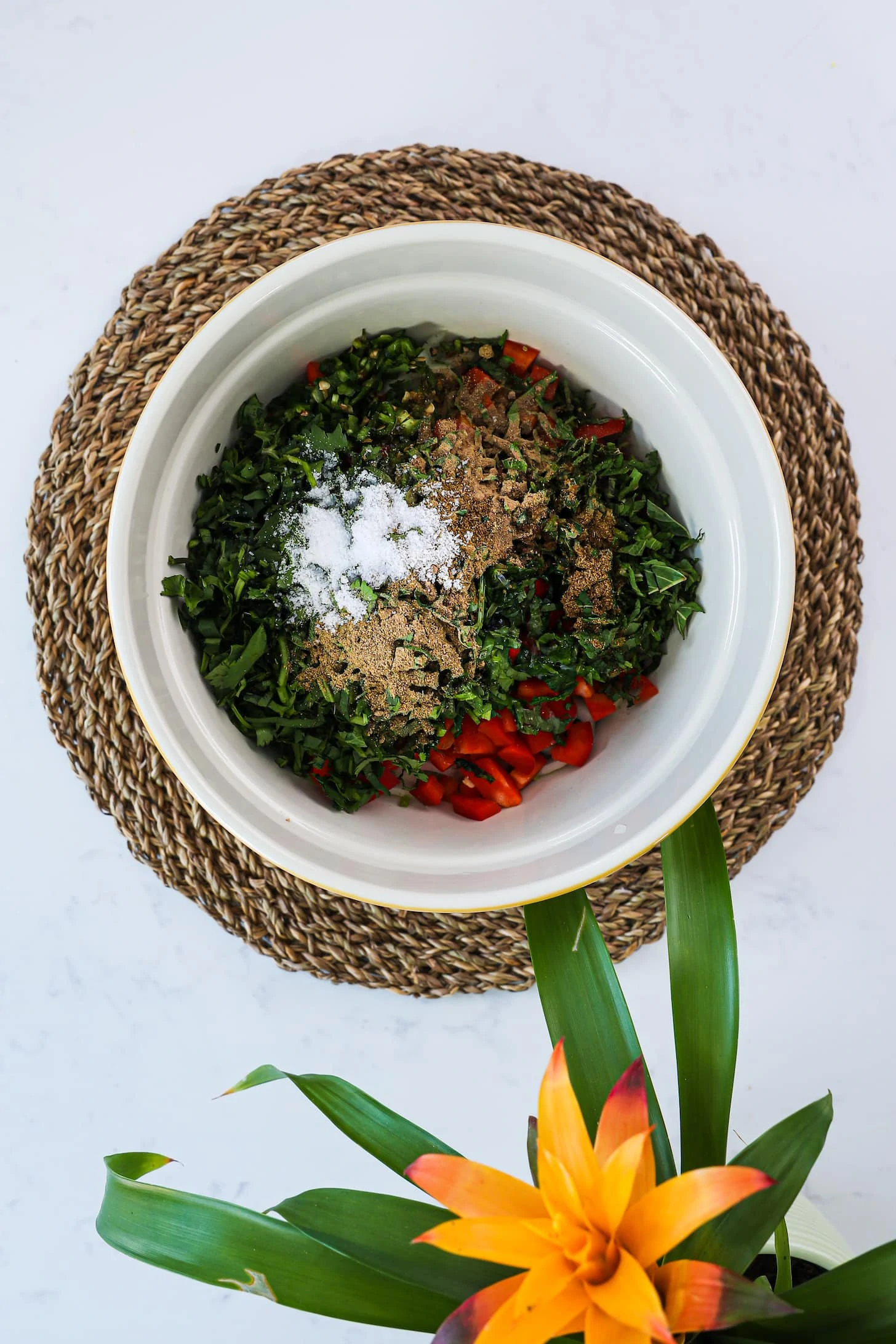 bowl of chopped herbs and peppers topped with spices and salt. Placed on top of straw place mat with a flowering plant in one corner.