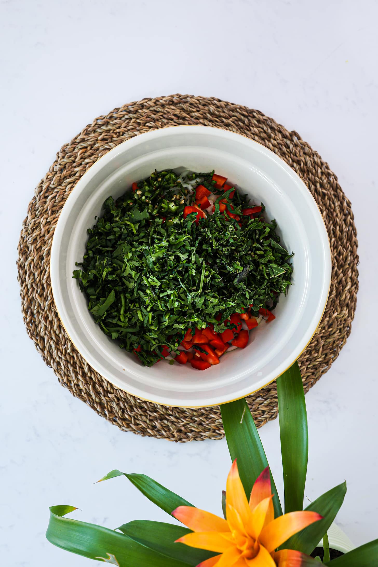 bowl of chopped herbs and peppers. Placed on top of straw place mat with a flowering plant in one corner.