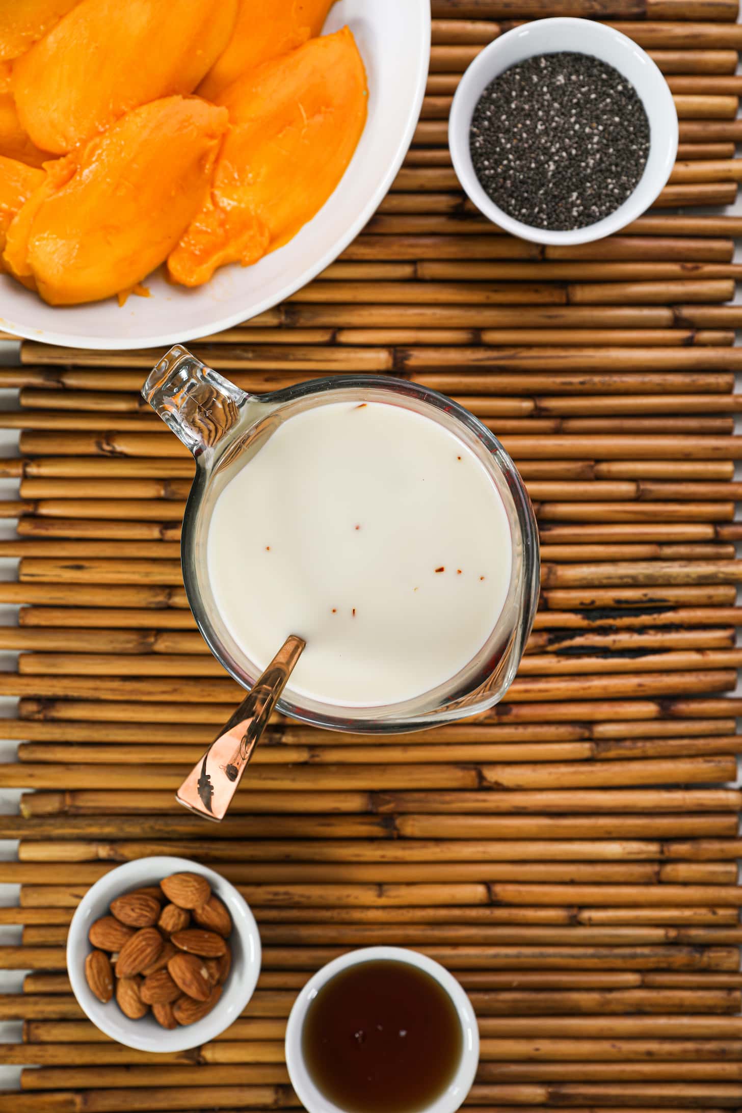 a jug of milk with a spoon in it surrounded by ramekins of ingredients including chia seeds, almonds, syrup and bowl of mango slices.