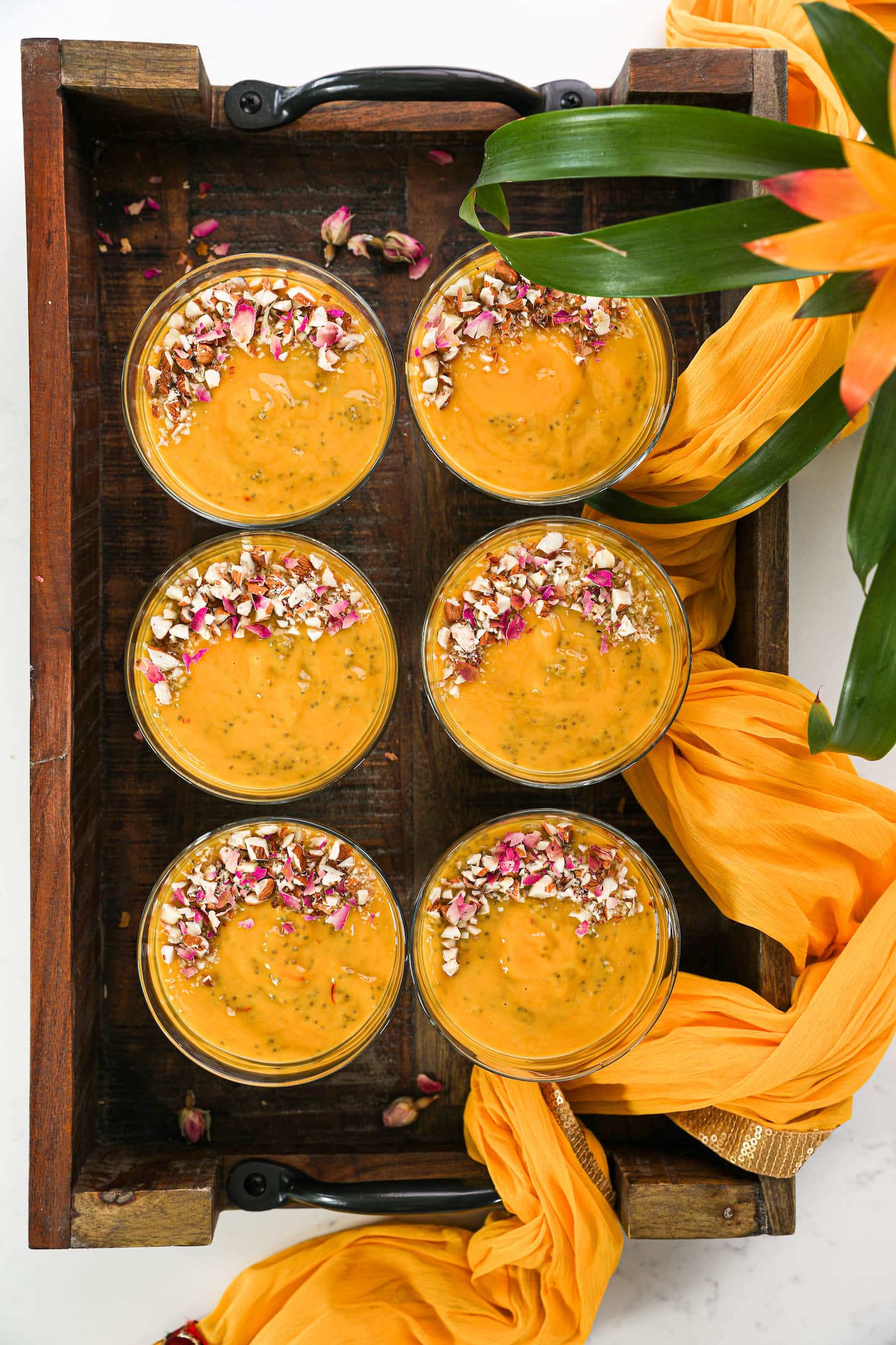 six serving bowls with a thick orange pudding in them garnished with nuts and dried roses placed placed inside a tray with a traditional scarf as decoration.