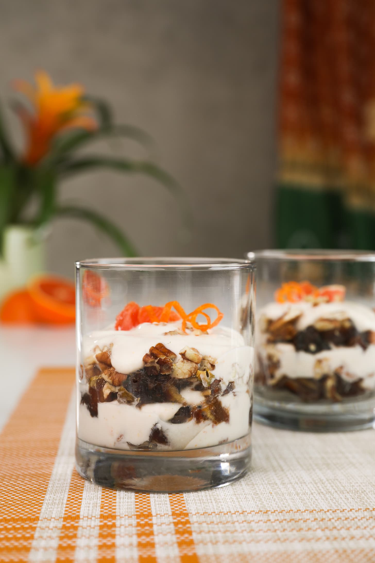 a glass of yogurt with layers of nuts topped with orange rind curls. Another glass with the same contents in seen in the background.