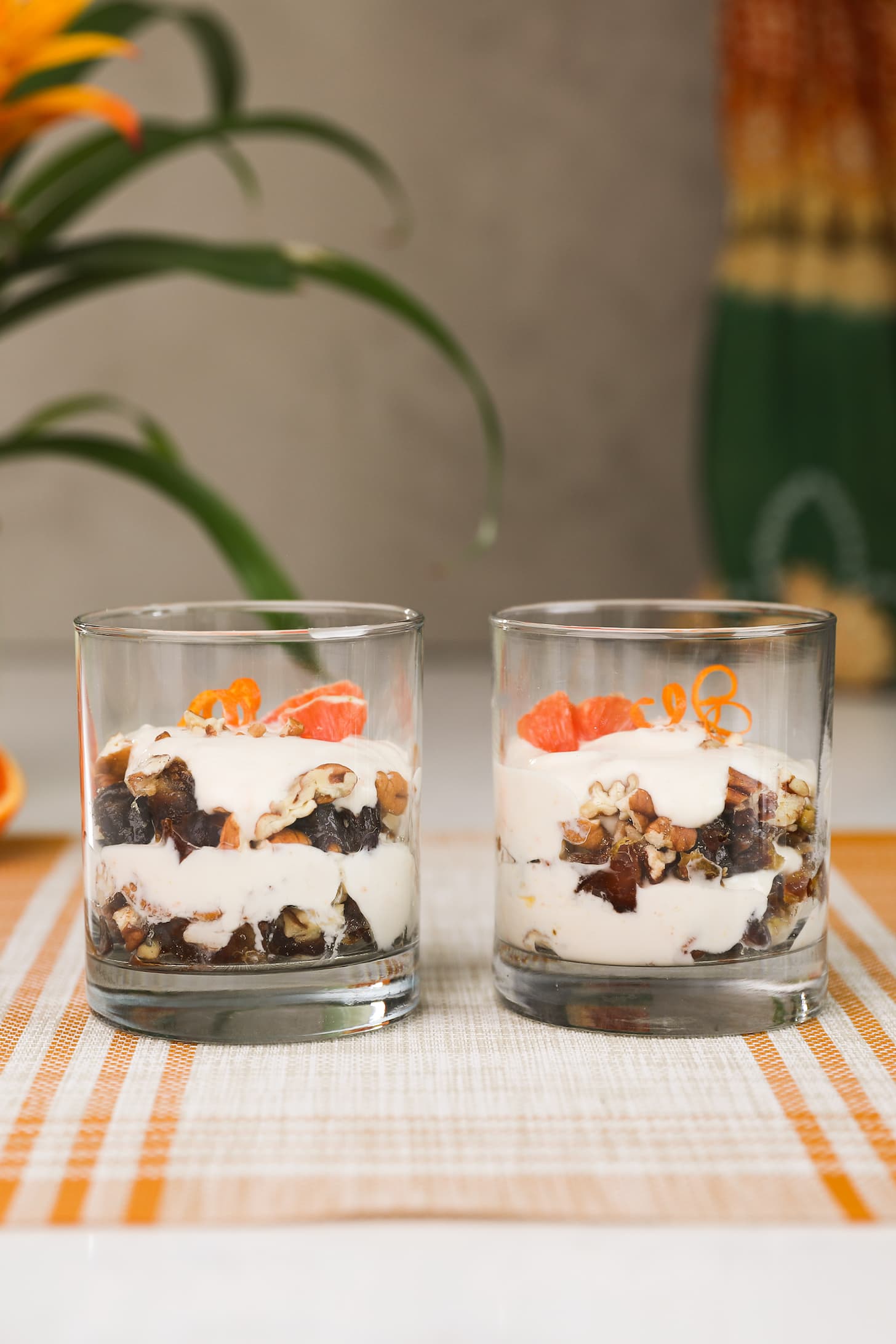 two glasses filled with layers of yogurt and nuts and topped with orange pieces and orange rind curls. There is also a plant in the background.