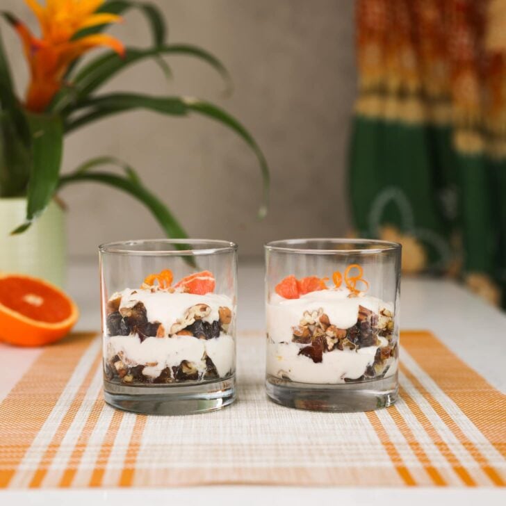 two glasses filled with layers of yogurt and nuts and topped with orange pieces and orange rind curls. There is also a plant in the background.