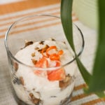 a glass filled with yogurt, topped with crushed nuts, orange rind curls and orange pieces.