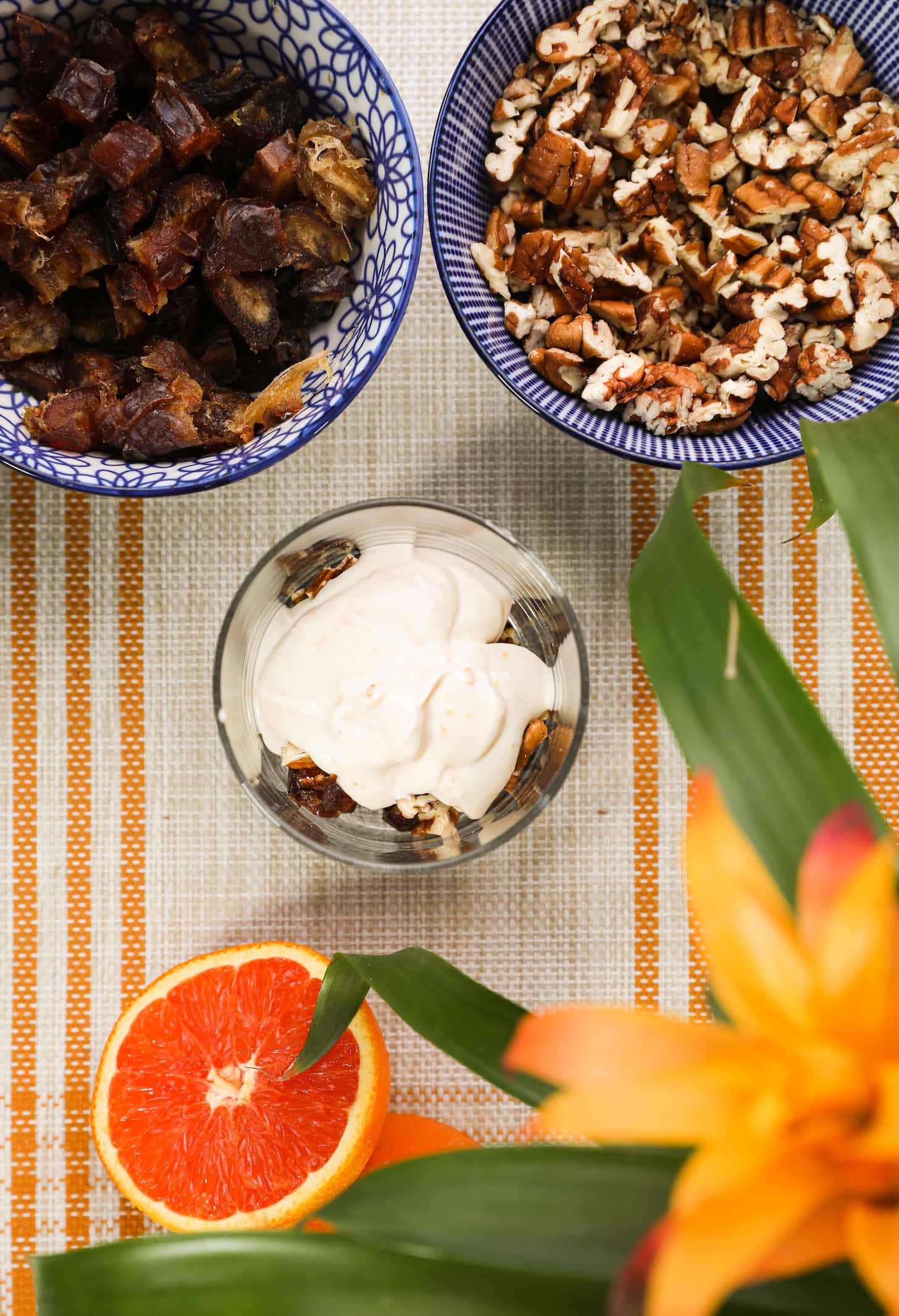 three different sized bowls filled with food: nuts, chopped dates and yogurt with half an orange and flowers in one corner.