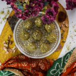 Eight brown nut covered balls on a gold printed plate on a yellow mat with flowers and an Indian decorative shawl on either side.