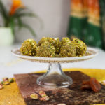 Nut covered brown balls on a gold printed decorative cake holder with a decorative shawl and a plant in the background.