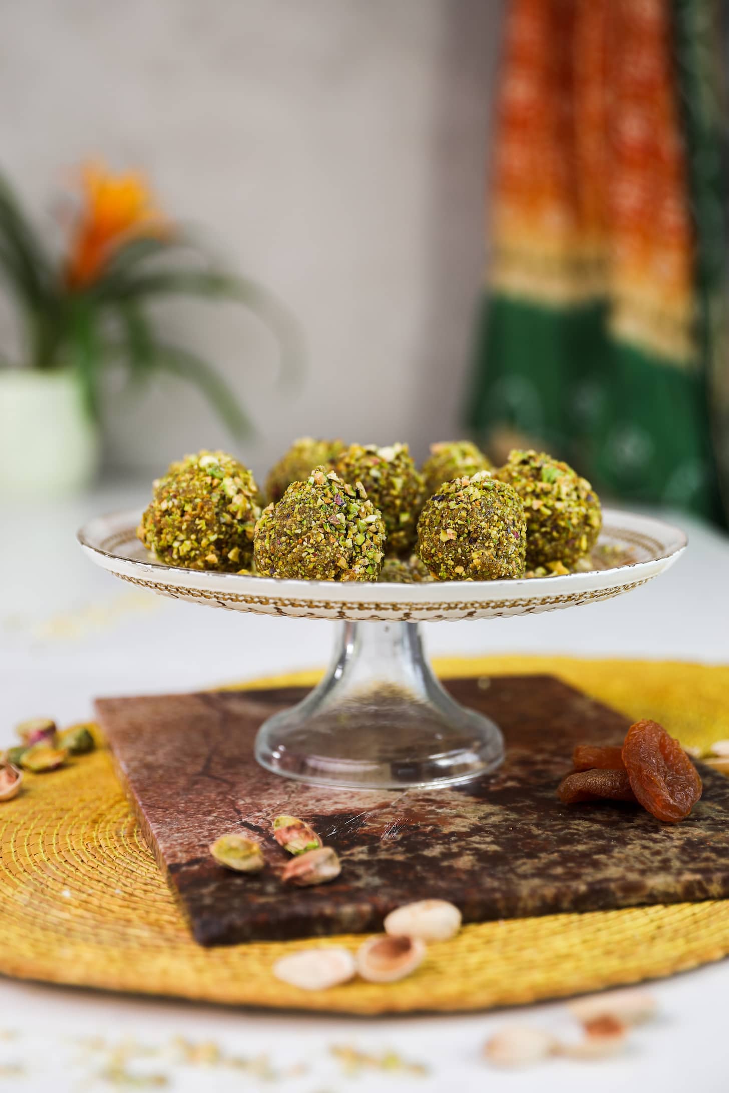 Nut covered brown balls on a gold printed decorative cake holder with a decorative shawl and a plant in the background.