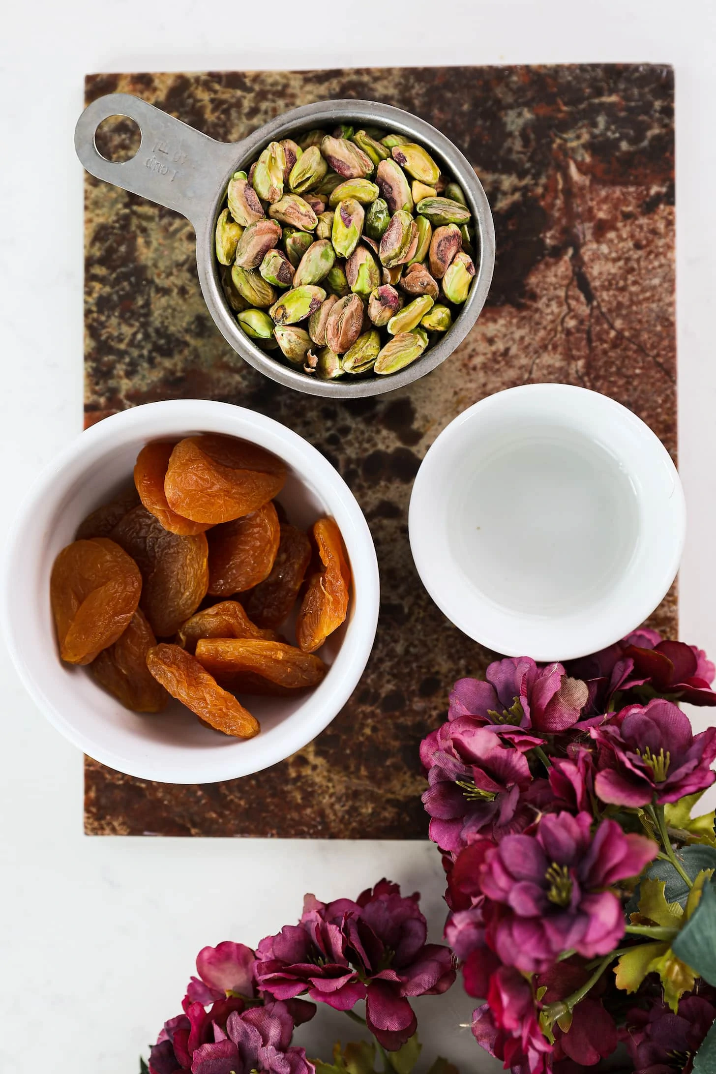 three different sized bowls holding pistachios, dried apricots and oil on a textured board with purple flowers in one corner.
